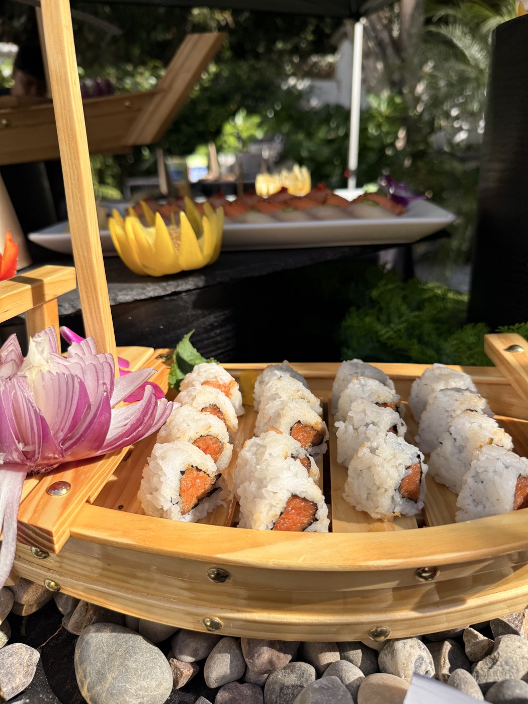 Bamboo boat of spicy tuna rolls with a carved red-onion lotus at the bow, in dappled garden light